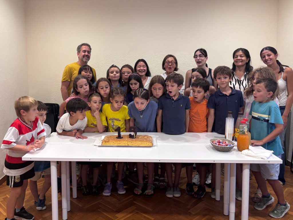 Alumnes bufant les espelmes del número 15 d’un pastís, amb el professorat al darrere, en una aula de l’Estudi Musical 143.